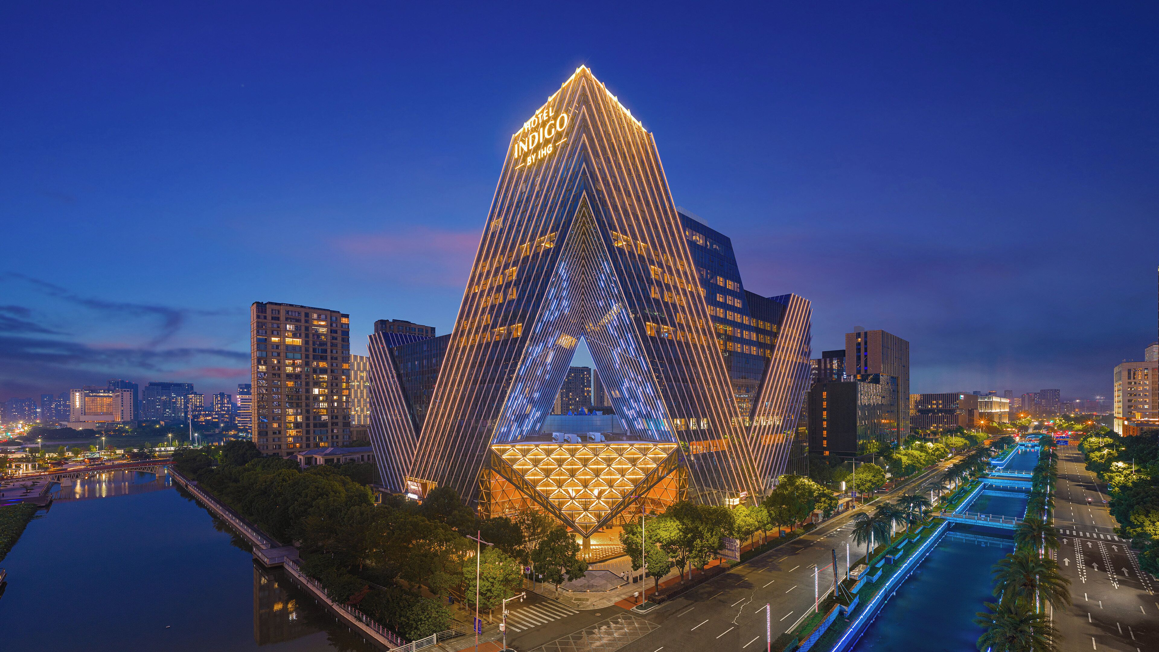 A modern hotel with a distinctive triangular facade, surrounded by a water body and urban skyline.