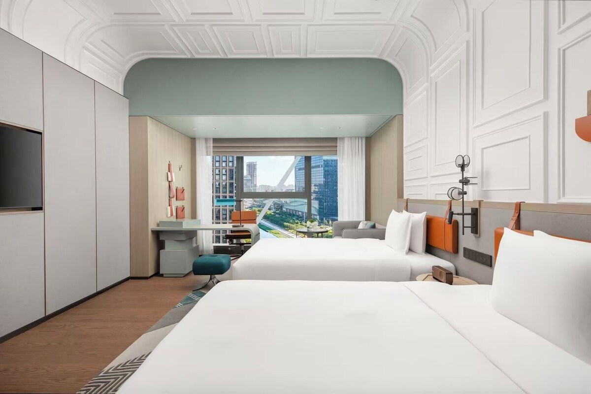 Habitación de hotel con dos camas, televisión, escritorio y vista a un paisaje urbano.