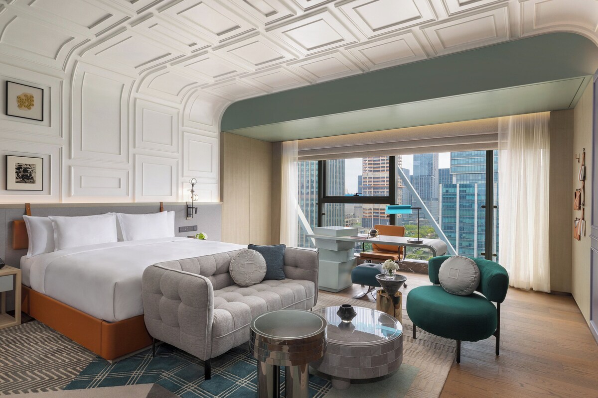 Una habitación de hotel moderna con una cama grande, un espacio de descanso con un sillón verde, una mesita y vistas a la ciudad.