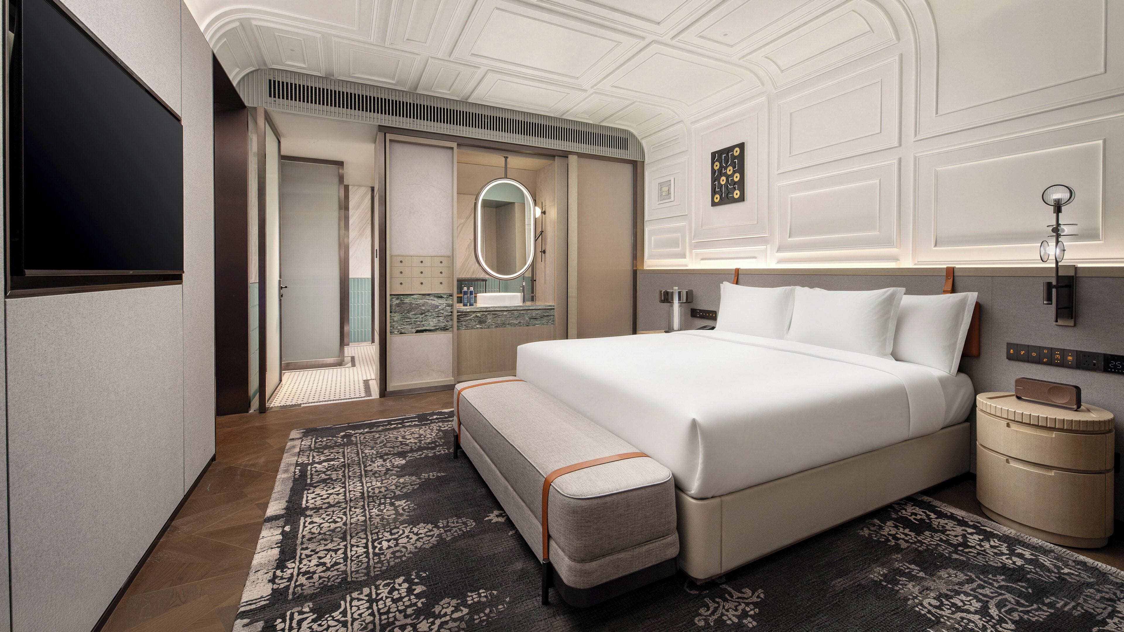 A modern hotel room with a large bed, a bedside table, a nightstand, a mirror, a wall clock, and a small bedside table with a lamp.