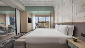 Habitación de hotel con una cama grande, un escritorio, una silla, un sofá, una mesita de noche y vistas a un paisaje urbano.