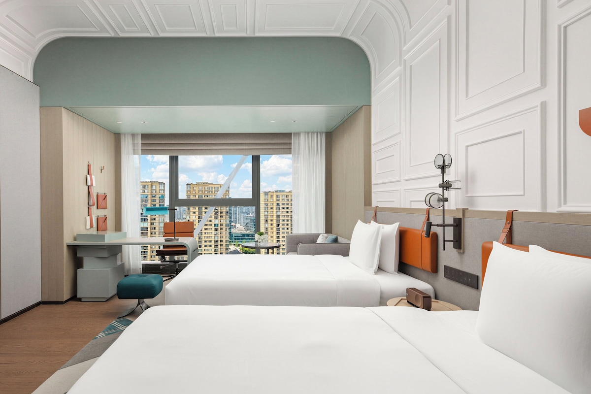 Habitación de hotel con dos camas, un escritorio, un televisor y vistas a la ciudad.