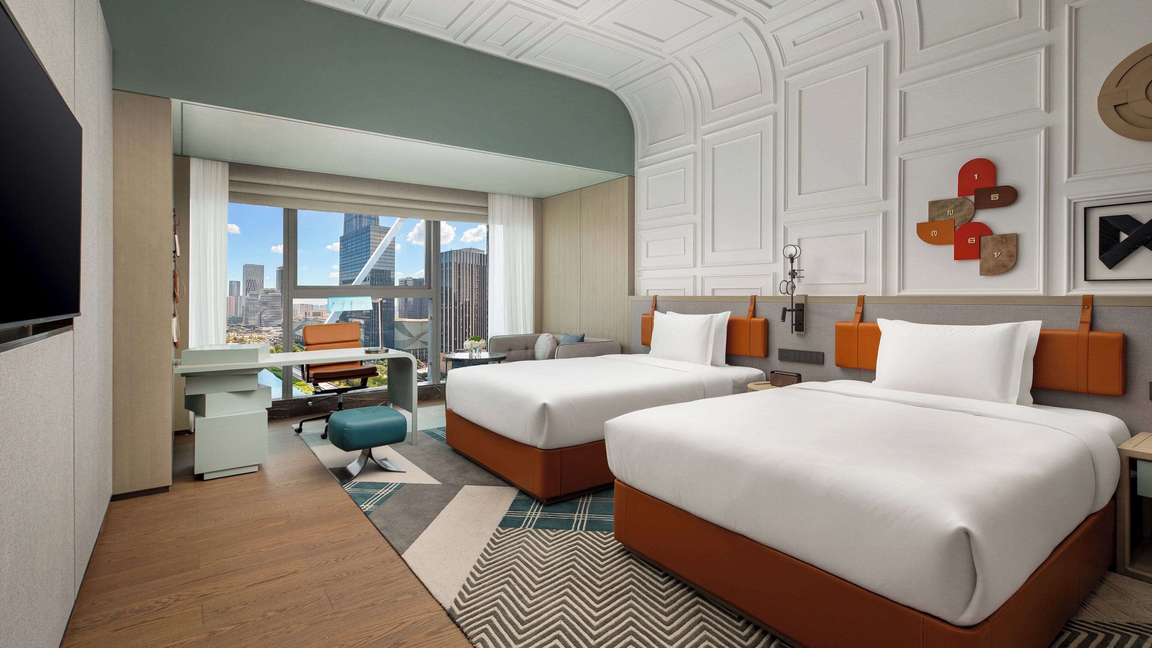 A hotel room with two beds, a desk, a chair, and a large window offering a city view.