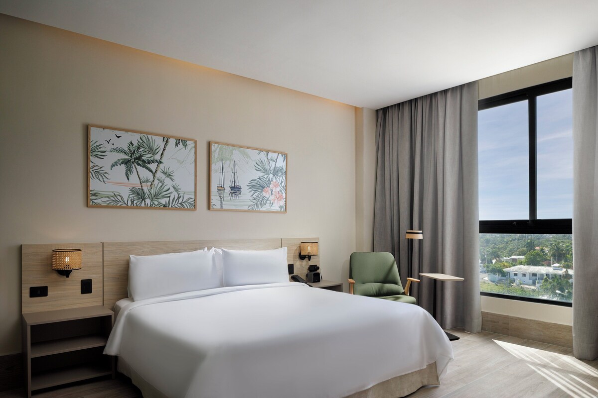 Ein Hotelzimmer mit einem großen Bett, zwei Wandlampen, einem grünen Sessel und einem Fenster mit Blick auf Gebäude.
