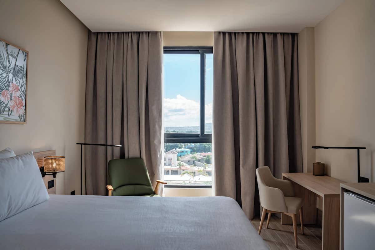 Ein Hotelzimmer mit Bett, Schreibtisch, Stuhl und Blick auf eine Stadtlandschaft durch große Fenster.