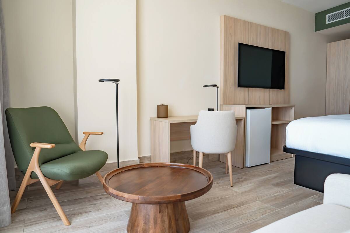 Ein modernes Hotelzimmer mit einem grünen Sessel, einem Holztisch, einem Schreibtisch mit Computer, einem Flachbildfernseher und einem Bett.