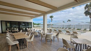 A modern outdoor dining area with tables and chairs, overlooking a pool and palm trees. - Holiday Inn Puerto Plata-Cofresi Bay Area By Ihg (Puerto Plata)