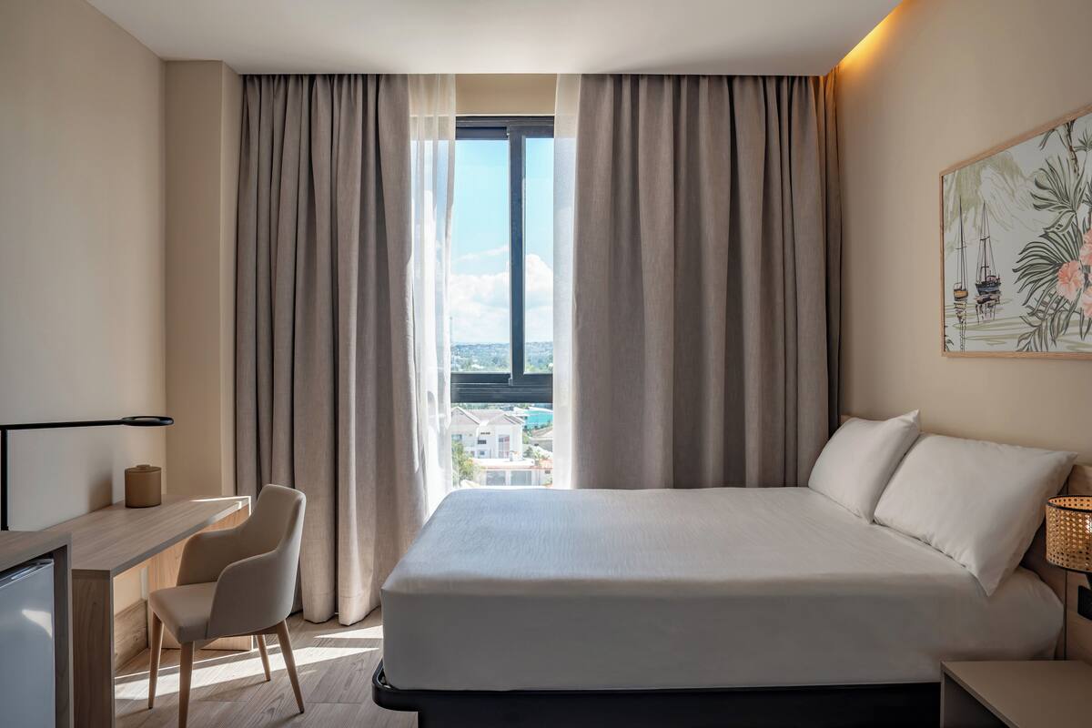 Ein Hotelzimmer mit einem Bett, einem Schreibtisch, einem Sessel und einem großen Fenster mit Vorhängen.