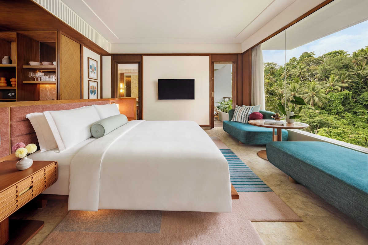 A modern hotel room with a large bed, a sofa, a TV, and a view of lush greenery.