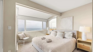 Une chambre avec un grand lit, une fenêtre donnant sur la mer, une chaise et une table de chevet avec une lampe.
