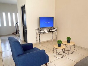 A living room with a blue sofa, a television on a white and black patterned table, and two small round tables with plants. - Sister Houses in private gated community with pool 5 min Airport and Consulate (Hermosillo)