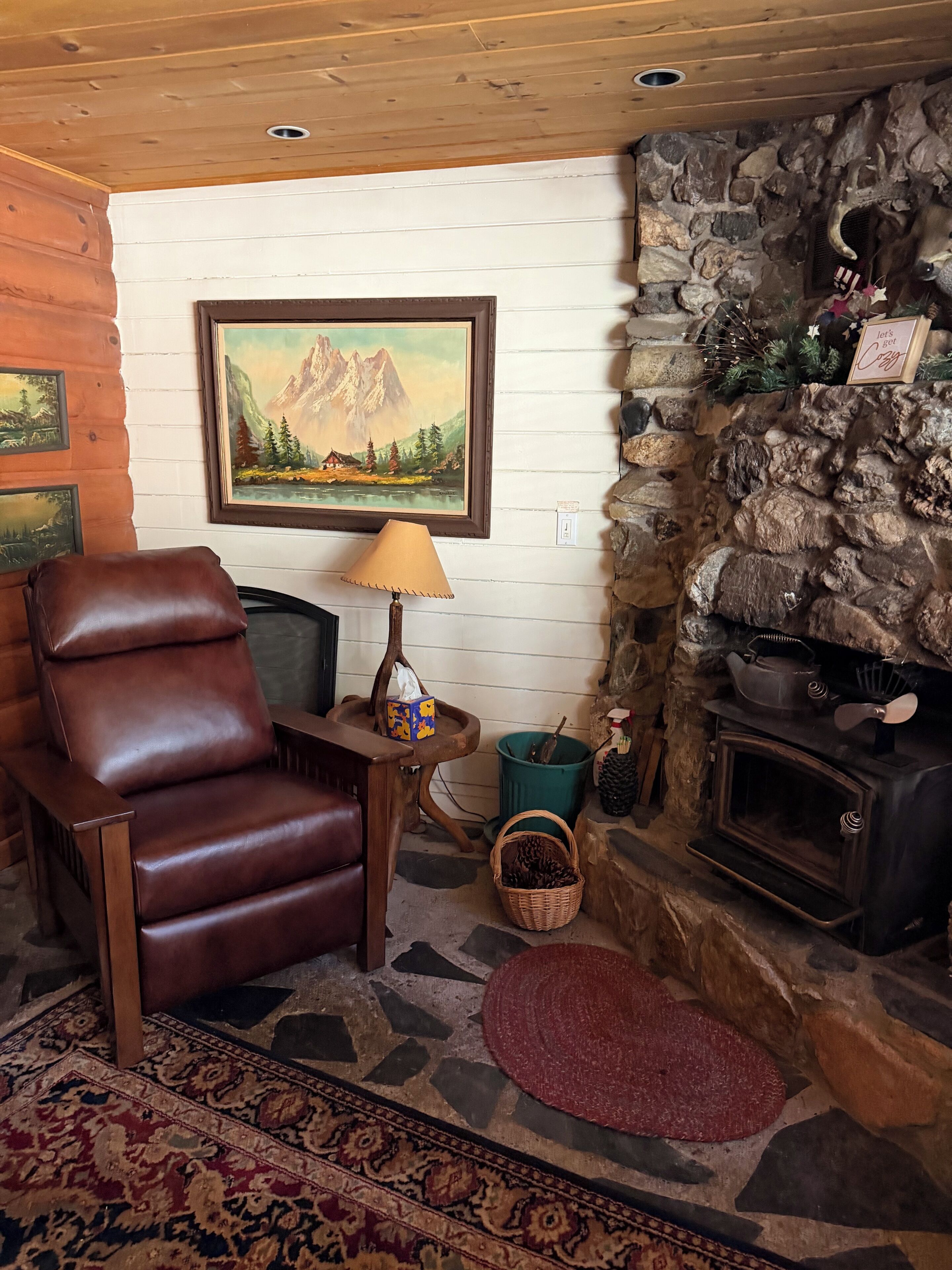 Interior d'una cabana de fusta amb una cadira de cuir, un quadre de muntanyes, una llar de pedra i una decoració rústica.