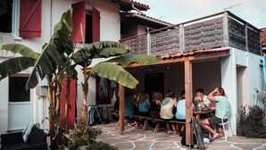 A group of people seated at an outdoor wooden table under a wooden pergola, with a surfboard leaning against the structure. - Surfbase Vieux Boucau (Vieux-Boucau-les-Bains)