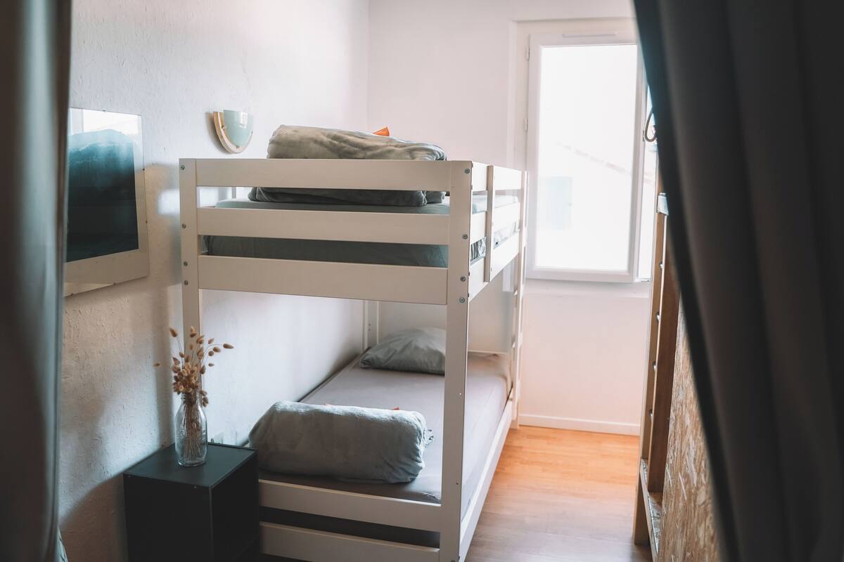 Ein Schlafraum mit Etagenbetten, einem Fenster, einem gerahmten Bild an der Wand und einer Nachttischlampe mit getrockneten Blumen.