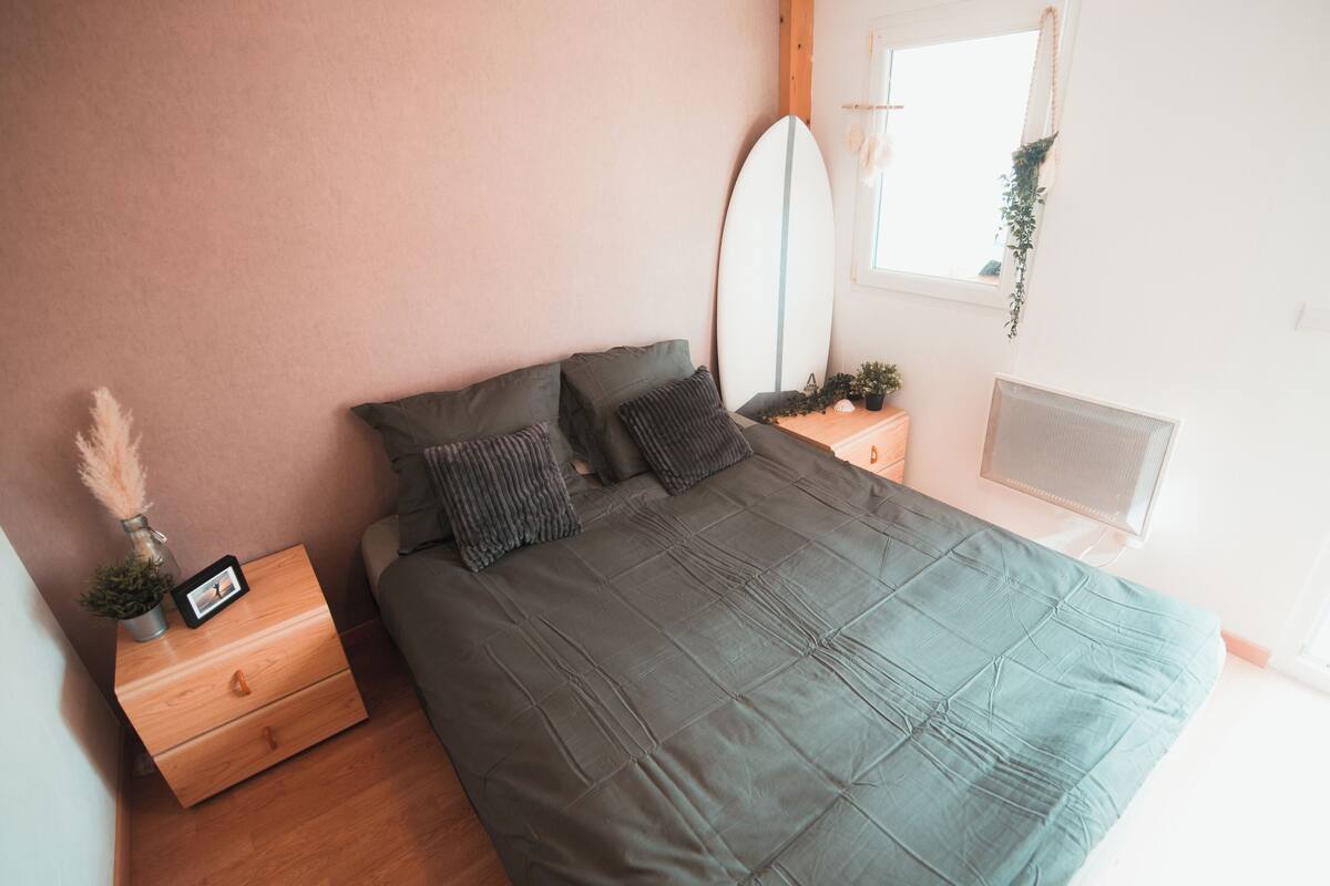 Ein Schlafzimmer mit Bett, Nachttisch, Schminktisch und Spiegel, sowie einem Fenster mit Pflanze.