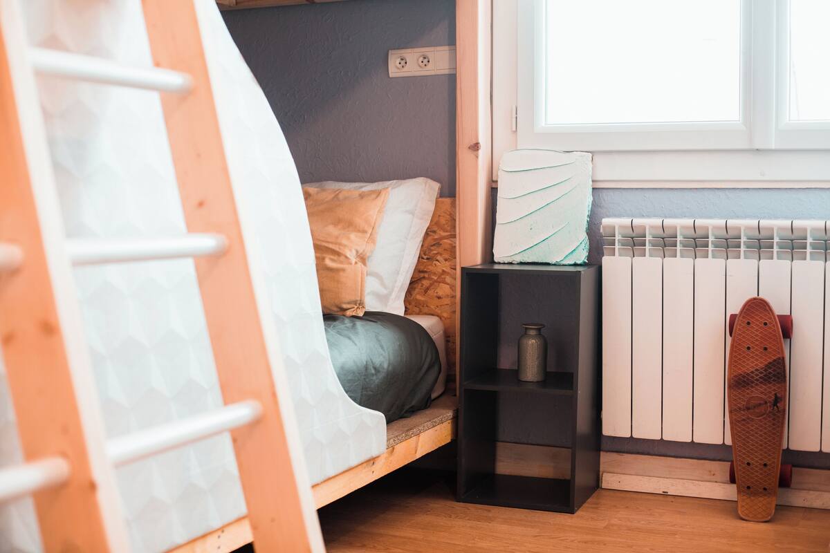 Ein Etagenbett mit Matratze und Kissen, eine kleine schwarze Ablage mit einer Flasche, ein Skateboard, das an einer Heizung anlehnt, und ein Fenster mit Jalousien.