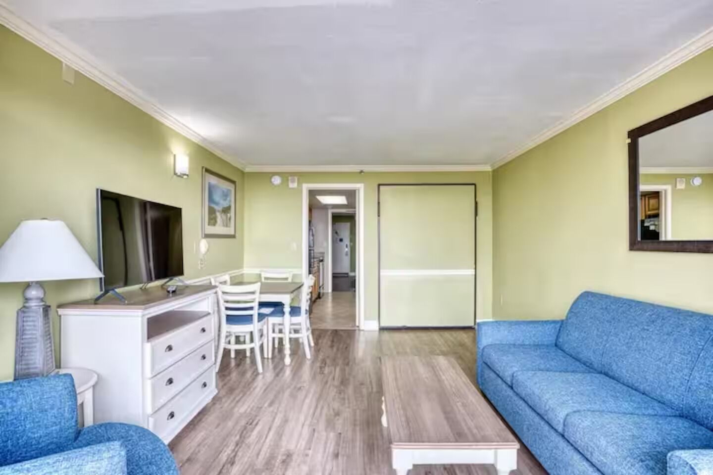 A room with a blue sofa, a white dresser, a desk with chairs, and a large mirror on the wall.