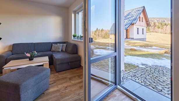Un salon moderne avec un canapé, un pouf et une table, de grandes fenêtres offrant une vue sur une maison et le paysage environnant.