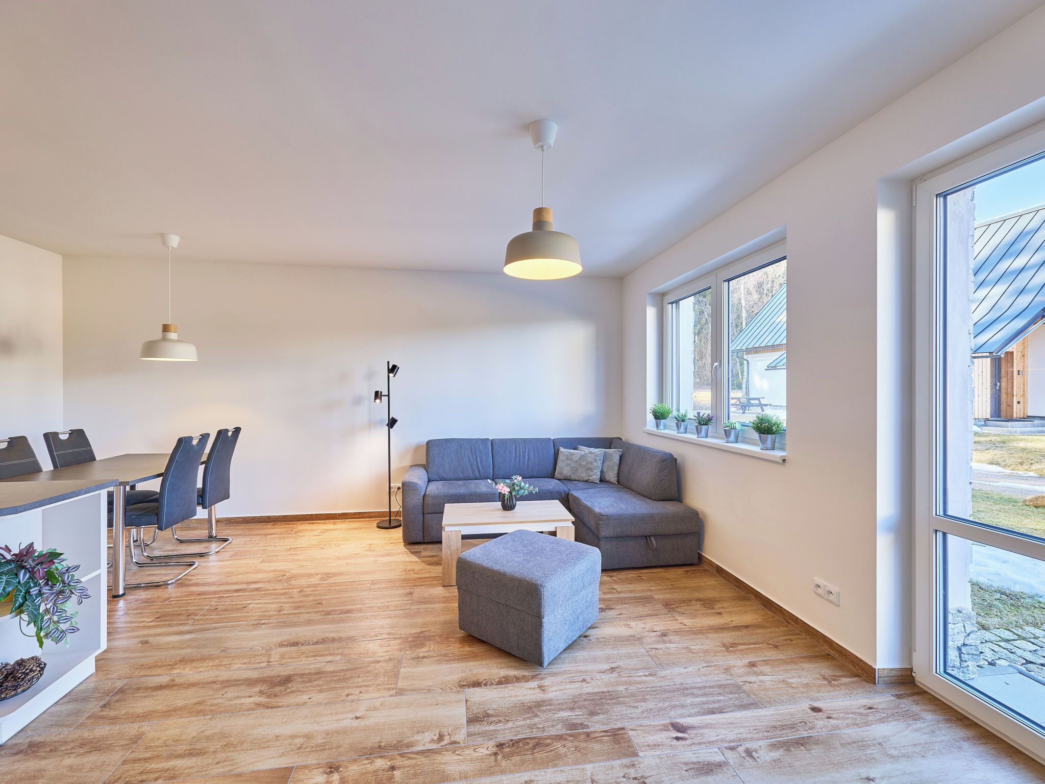 A modern living room with a sofa, ottoman, dining table, chairs, pendant lights, and large windows.
