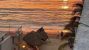 Une vue sur le coucher de soleil au-dessus de l’océan, avec une plage et une structure surmontée d’un toit de chaume.