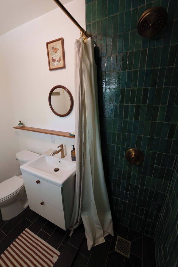 Een badkamer met een wit wastafelmeubel, toilet en douche met een wand betegeld in groene tegels, plus een lijst met een afbeelding aan de muur.
