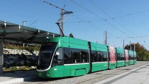 A green tram at a station with overhead wires, a covered waiting area, and a stone wall in the background. - Studio near Basel Airport RM1142 (Saint-Louis)