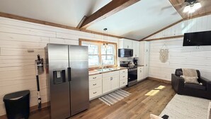 A compact kitchen with a stainless steel refrigerator, a microwave, and a stove. There is a window with a view of trees, a ceiling fan, and a black leather chair with a cushion. - Cozy 1-bedroom cabin in the Hocking Hills of Ohio (Londonderry)