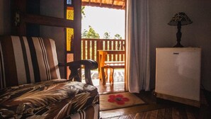Una habitación con una cama, una silla, una mesita y un refrigerador. Fuera de la puerta, hay un banco de madera.
