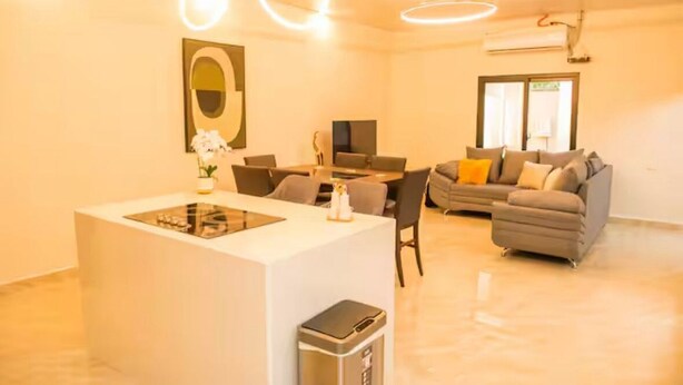 A modern living room with a sofa, a dining table, and a small kitchen area with a sink and a trash bin. - Villa Idalia (Comayagua)