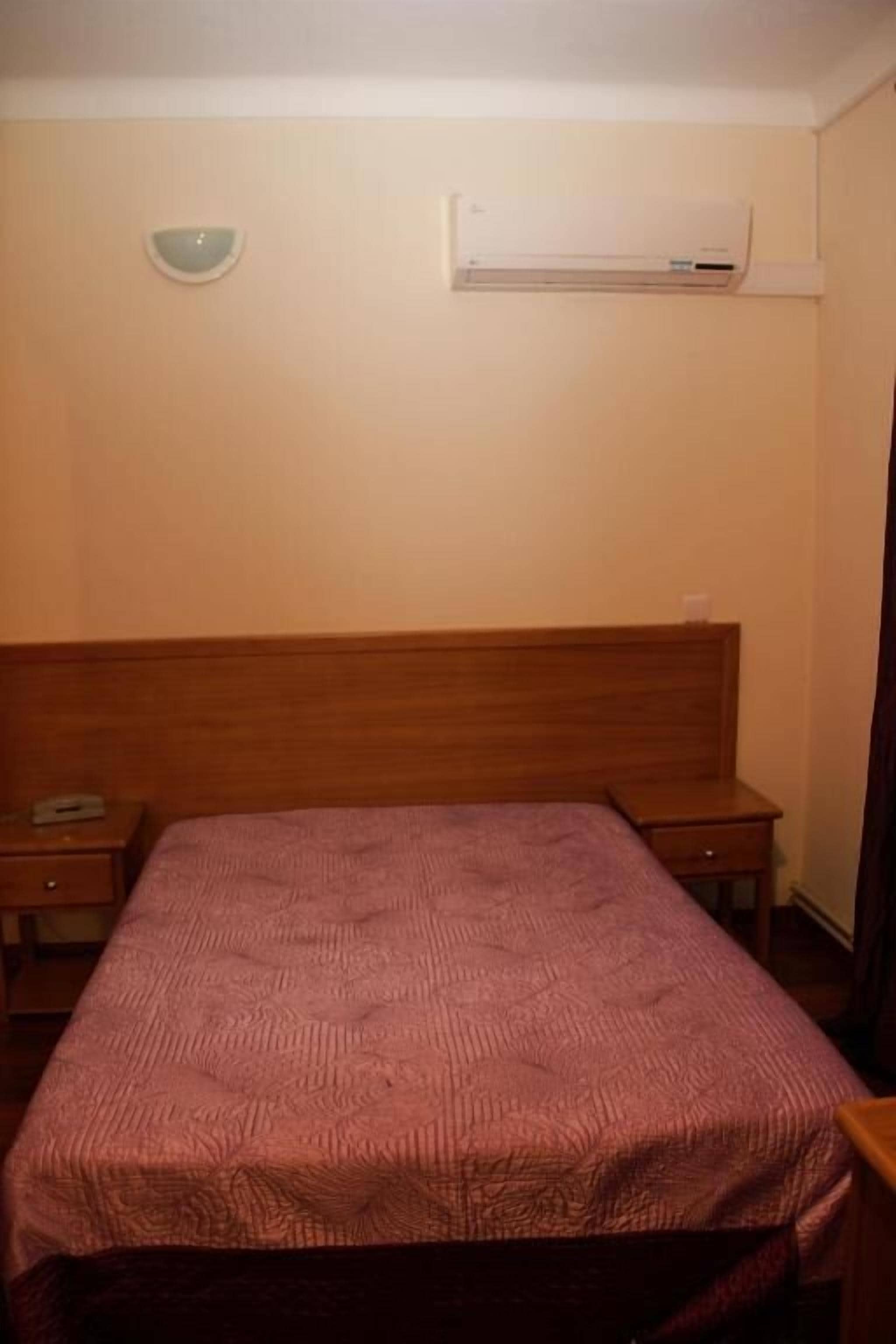 A room with a bed, a bedside table, a nightstand, an air conditioning unit, and a wall-mounted light fixture.