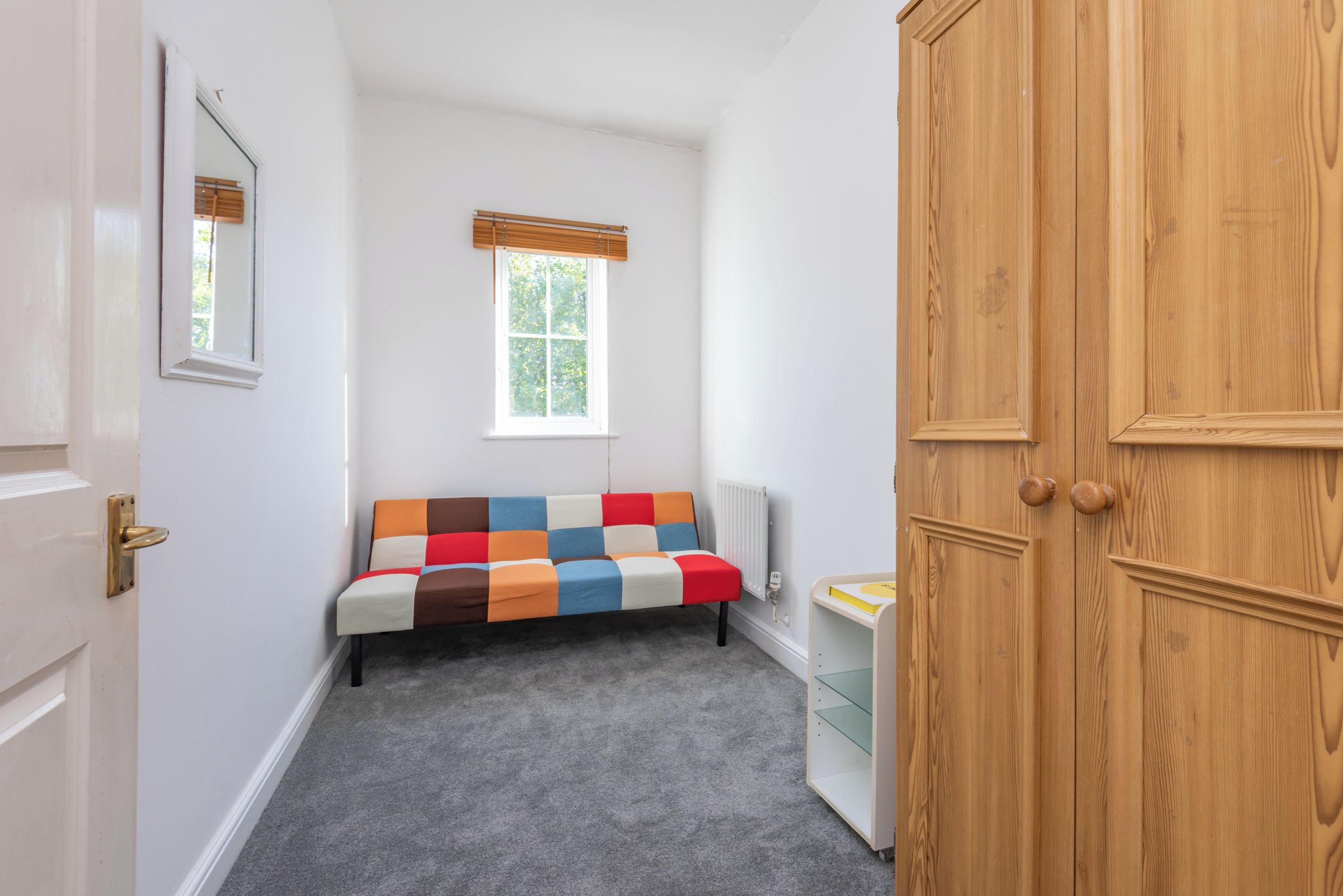 A small room with a grey carpet, a colorful sofa, a radiator, and a wooden door.