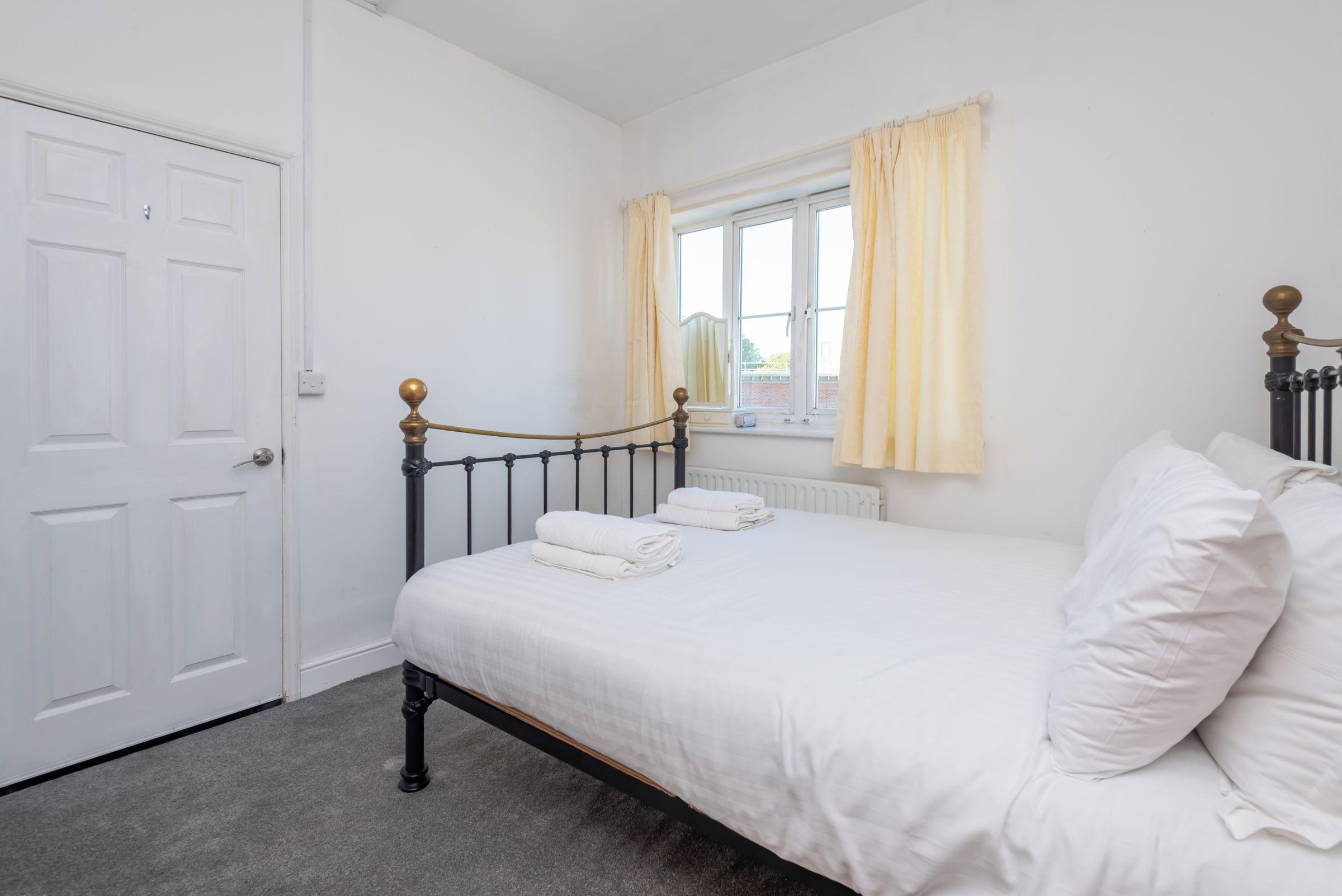 A bedroom with a single bed, white bedding, a radiator, a window with curtains, and a door.