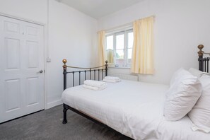 Quarto com uma cama de solteiro, roupa de cama branca, radiador, janela com cortinas e porta.