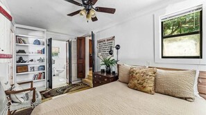 A bedroom with a bed, a ceiling fan, a bookshelf, a window with a view, and a door leading to another room.