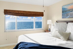 A hotel room with a large window overlooking the ocean, a bed with white linens, a nightstand with a lamp, and a painting on the wall. - Beachfront Condo on Bonita Beach  - SeaGlass Shores (Bonita Springs)