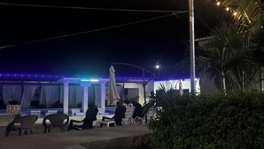 Un espacio al aire libre con sillas y mesas, luces colgantes y un edificio con fachada iluminada en azul al fondo.