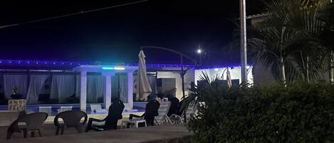 An outdoor seating area with chairs and tables, string lights overhead, and a building with a blue-lit facade in the background.