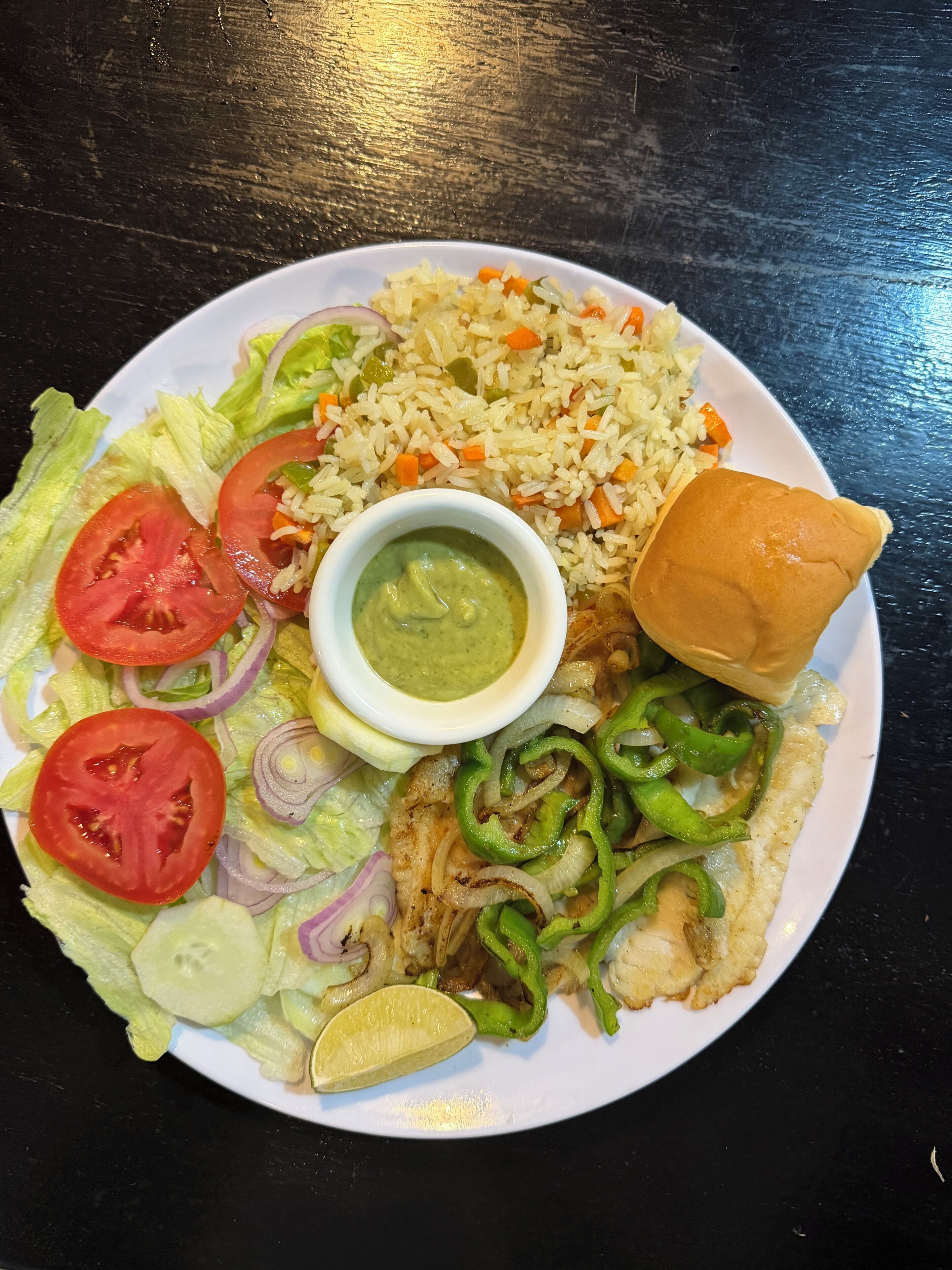 A plate with rice, vegetables, a piece of meat, a bun, and a small bowl of sauce.