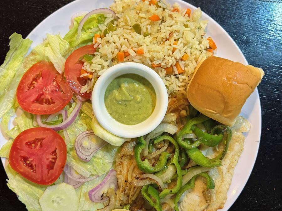 A plate with rice, vegetables, a piece of meat, a bun, and a small bowl of sauce.