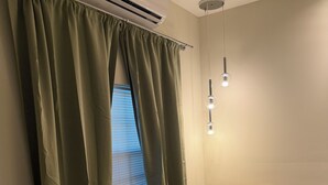 Una habitación con una ventana cubierta por cortinas verdes, un sillón color beige con un cojín y una lámpara colgante.