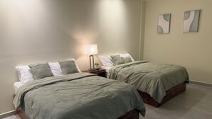 Un dormitorio con dos camas, cada una con cabecero de madera y colcha verde. Entre las camas hay una mesita de noche con una lámpara y una pequeña planta en maceta. Las paredes son blancas y el suelo está cubierto de baldosas.