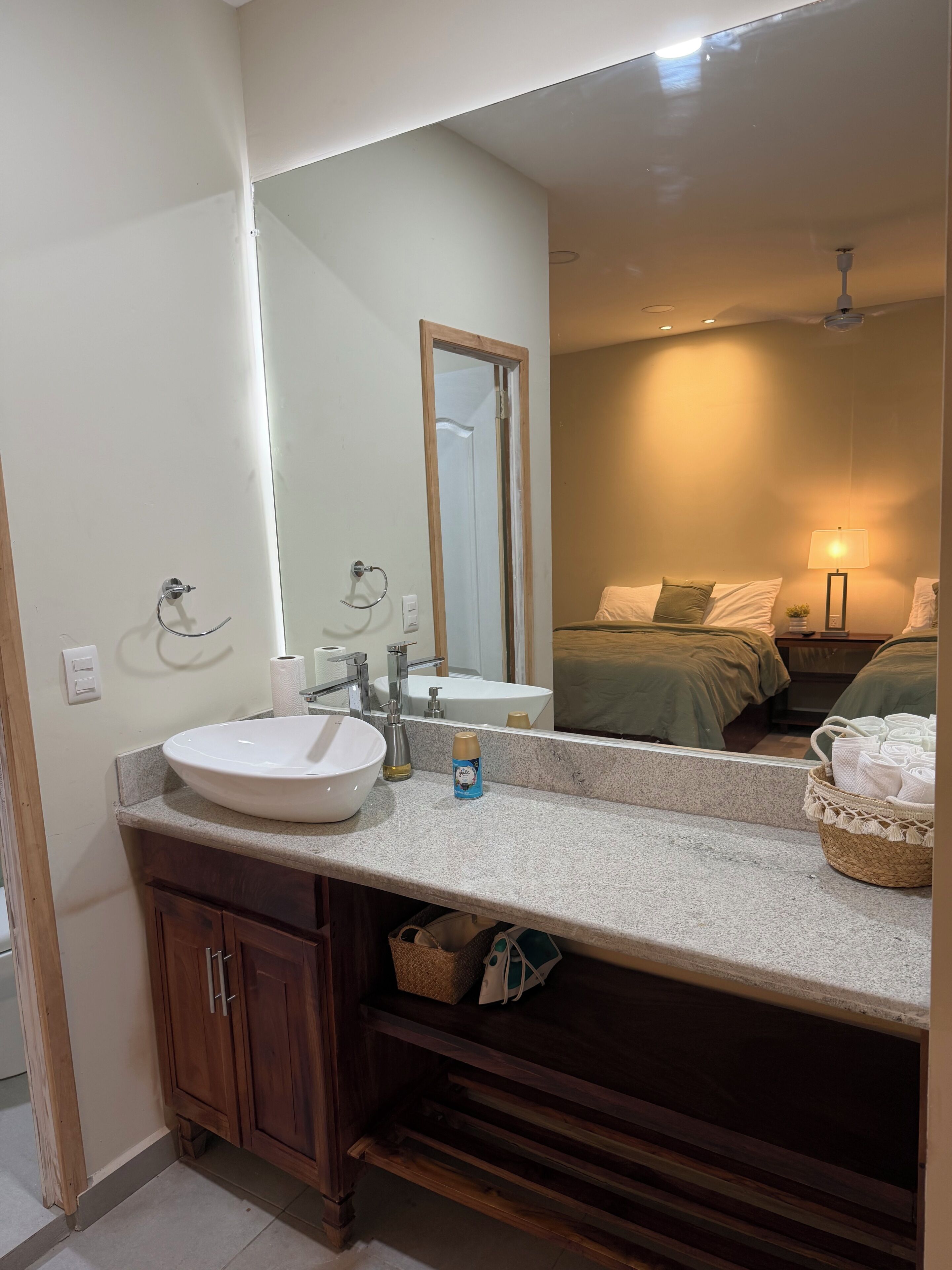 A bathroom with a sink, mirror, and a bed visible in the reflection.
