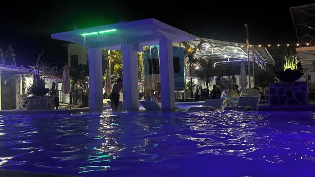 Un área de piscina nocturna con zona de estar cubierta y palmeras.
