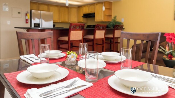 A dining table set with plates, glasses, and cutlery, with a kitchen in the background. - One Bedroom Suite (Freeport)