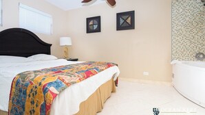 A bedroom with a bed, a ceiling fan, a nightstand with a lamp, two framed artworks, and a bathroom with a tiled shower wall. - One Bedroom Suite (Freeport)