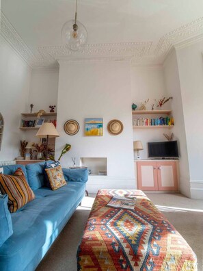 A living room with a blue sofa, a patterned ottoman, a television, and bookshelves.