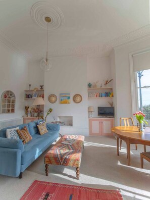 Una sala de estar con un sofá azul, un reposapiés estampado, una mesa de comedor de madera con sillas, un televisor y una estantería.