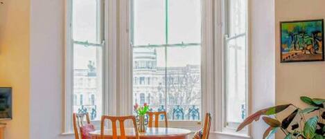 Un salón luminoso con un sofá azul, una mesa de comedor con sillas, una alfombra roja y un ventanal con vistas a edificios.