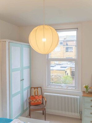 A room with a large window, a radiator, a wardrobe, a chair, and a hanging light fixture.