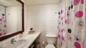 Ein Badezimmer mit weißem Waschbecken, Spiegel und Toilette. Ein blumiges Duschvorhang und ein kleiner Korb mit einer Blume stehen auf der Ablagefläche.